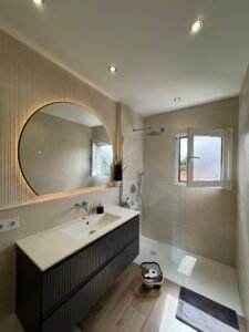 Baño de diseño contemporáneo en tonos claros, con gran espejo circular con retroiluminación y mampara de cristal, creando un espacio luminoso, elegante y perfectamente optimizado